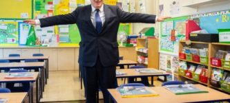 UK+Prime+Minister+Boris+Johnson+demonstrates+how+schools+are+Covid-safe+by+holding+two+arms+over+three+chairs